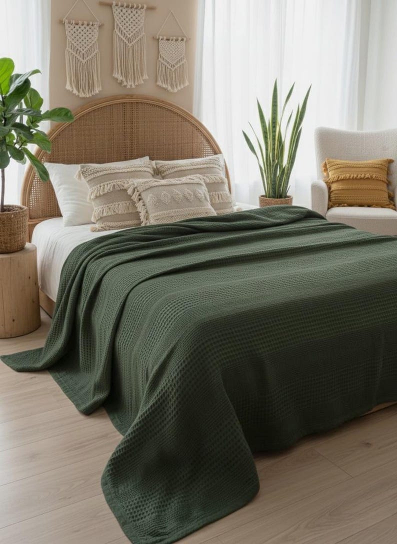 May include: A dark olive green waffle-weave blanket draped over a bed. The blanket features a textured grid pattern across its entire surface, providing a uniform appearance. It is positioned to cover the mattress, extending down to the floor.