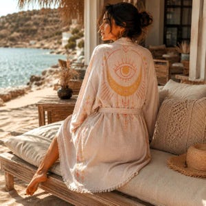 May include: A beige kimono robe featuring a large orange and yellow graphic print on the back depicting an eye and a crescent moon. The garment has long sleeves, a tie waist belt, and a fringed hemline, crafted from a textured fabric.