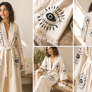 May include: Cream-colored robe with a repeating black eye design. The robe has long sleeves, a tie waist, and a flowing silhouette. The eye motif is printed on the front, sleeves, and back. The fabric appears to be lightweight and textured, suitable for lounging.