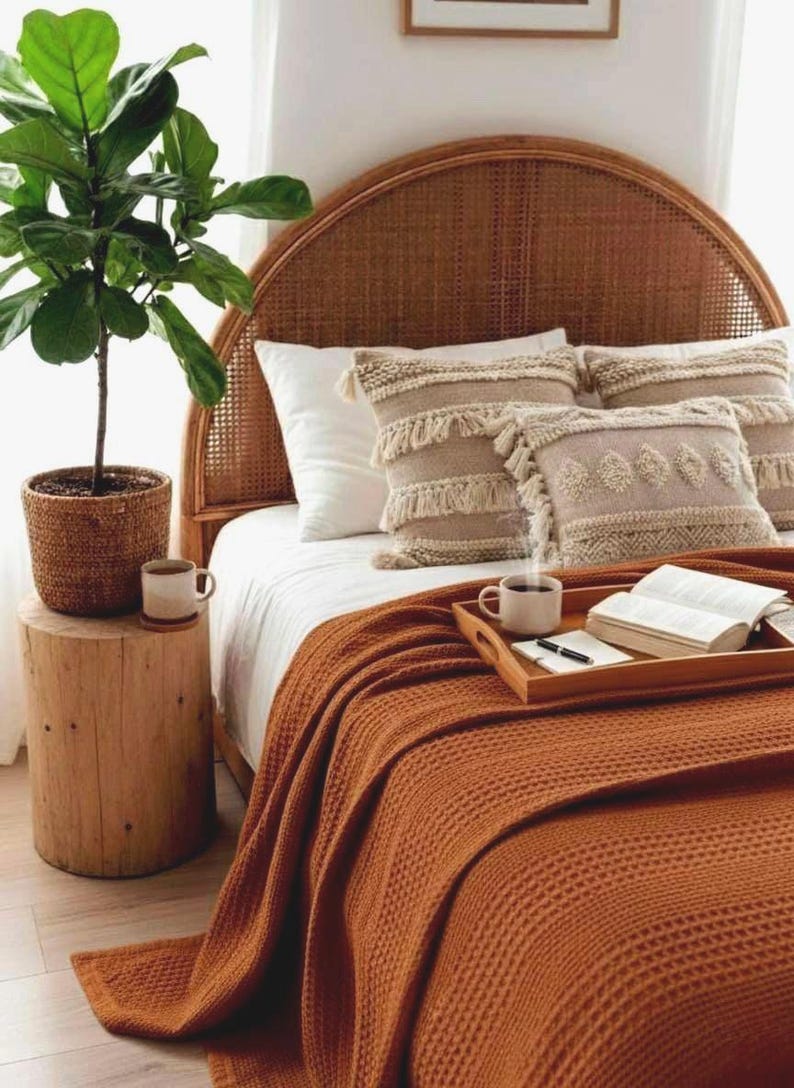 May include: A cozy bedroom scene featuring a bed with a woven headboard, white bedding, and decorative pillows. A rust-colored waffle-knit blanket is draped over the bed. A potted plant and a wooden side table with a coffee cup complete the warm, inviting aesthetic.