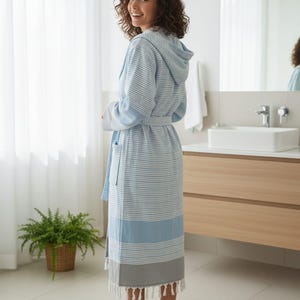 May include: A hooded bathrobe featuring a pattern of thin horizontal white and light blue stripes. The robe includes a matching tie belt at the waist, long sleeves with solid light blue cuffs, and a wide grey hem finished with white fringe tassels.