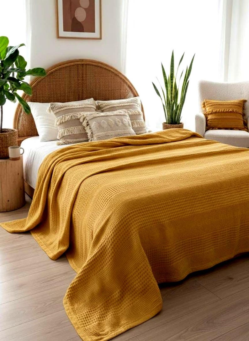 May include: A mustard yellow waffle-weave bedspread draped over a bed with a rattan headboard. The bed features two beige decorative pillows with fringe and a white pillow. A matching mustard yellow pillow rests on a nearby armchair.
