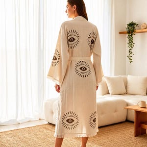 May include: A beige linen robe with a repeating eye print in dark brown. The robe has a matching belt and fringed edges on the sleeves and hem. The model is wearing the robe in a well-lit room.