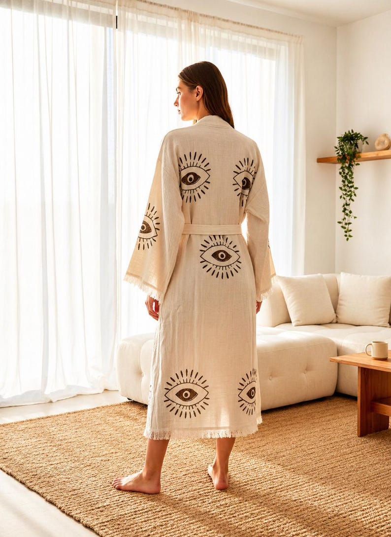 May include: A beige linen robe with a repeating eye print in dark brown. The robe has a matching belt and fringed edges on the sleeves and hem. The model is wearing the robe in a well-lit room.