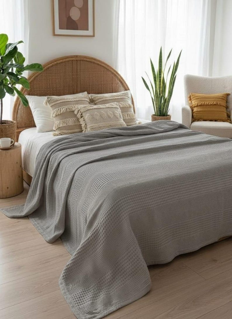 May include: A light gray waffle-weave blanket draped over a bed. The bed features a rounded rattan headboard, white pillows, and two decorative beige throw pillows with textured patterns. A green plant sits in a woven basket nearby.