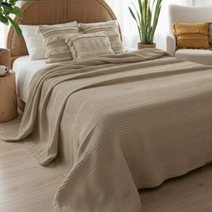 May include: A beige waffle-weave bedspread draped over a bed with a rattan headboard. The bed features two rectangular throw pillows with fringe and textured patterns, alongside plain white pillows, creating a layered bedding arrangement.