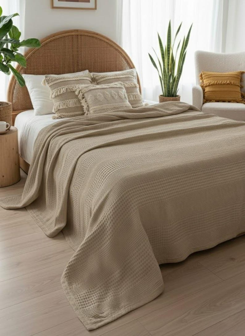 May include: A beige waffle-weave bedspread draped over a bed with a rattan headboard. The bed features two rectangular throw pillows with fringe and textured patterns, alongside plain white pillows, creating a layered bedding arrangement.