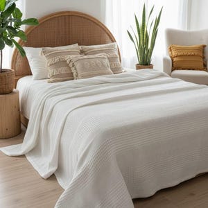 May include: A white waffle-weave bedspread draped over a bed with a rounded rattan headboard. The bed features two plain white pillows and two beige decorative pillows with fringe and textured patterns, creating a layered bedding arrangement.