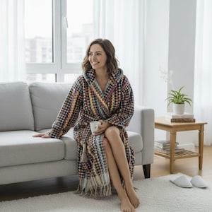 May include: A woman wearing a hooded, multi-coloured patterned robe, sitting on a grey sofa. The robe has a fringed hem and is made of a soft, textured material. She holds a white mug. The room has a large window and a light-coloured rug.