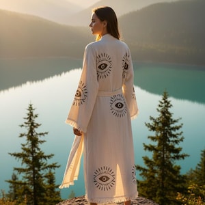 May include: A cream-colored robe with a repeating eye design in black. The robe has long sleeves, a belt, and fringe along the bottom. The person is standing on a rock, overlooking a lake and mountains.