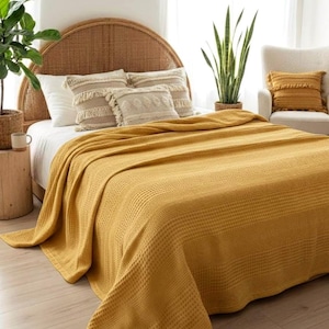 Puede incluir: Una manta de punto de gofre amarillo mostaza sobre una cama. La cama tiene almohadas decorativas y un cabecero tejido. Una almohada a juego está en una silla. La habitación está decorada con plantas y una obra de arte enmarcada.