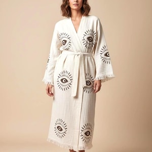 May include: Cream-colored robe with a repeating eye design in dark brown. The robe has a tie belt, long sleeves, and fringe trim. The fabric appears to be a lightweight cotton or linen. The robe is knee-length.