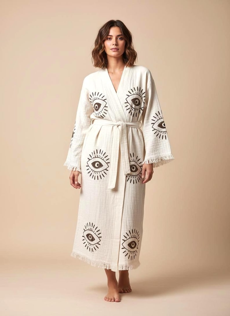 May include: Cream-colored robe with a repeating eye design in dark brown. The robe has a tie belt, long sleeves, and fringe trim. The fabric appears to be a lightweight cotton or linen. The robe is knee-length.