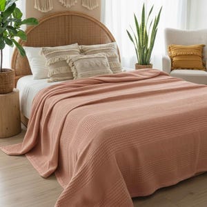 May include: A solid dusty rose colored blanket with a waffle weave texture draped over a bed. The blanket features a grid-like pattern across its entire surface and hangs over the edges of the mattress, covering the white bed sheets underneath.