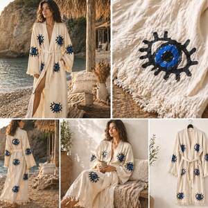 May include: A cream-colored robe with a belt and wide sleeves, adorned with embroidered blue and black eye motifs. The robe has a fringed hem and is made of a lightweight fabric. The robe is shown in multiple settings, including a beach and a room.