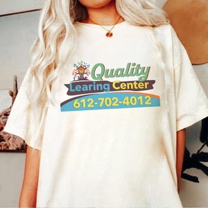 May include: Cream-colored t-shirt featuring the words "Quality Learning Center" in green, blue, and yellow. The design includes a treehouse graphic and a phone number. The shirt is a casual, short-sleeved style.