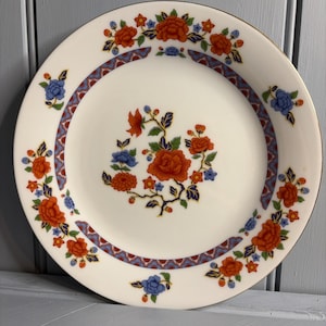 May include: White ceramic plate with a gold rim and a floral design. The plate features a central cluster of orange flowers with blue accents and a border of orange and blue floral patterns. The plate is a decorative piece.