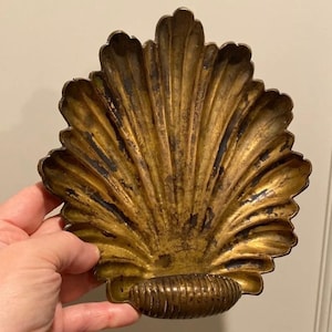 May include: A vintage, gold-toned, shell-shaped wall sconce. The sconce has a detailed, textured surface with a ribbed base. The shell design features radiating lines and a slightly distressed finish, adding to its antique charm.