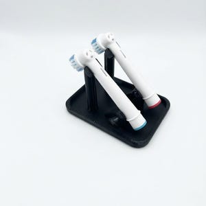 May include: A black plastic stand holding two white electric toothbrush heads. Each head features blue and white bristles, with one base ring colored light blue and the other base ring colored pink, positioned upright on the rectangular base.