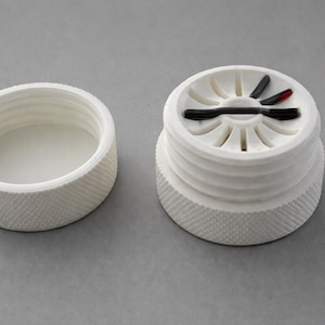 May include: A white bottle stopper with a textured exterior and a separate, matching cap. The stopper has a unique design with black and red accents. The cap is round with a textured, ridged pattern.