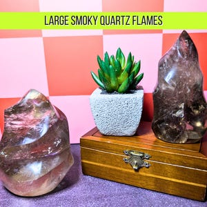 May include: Two large smoky quartz flame sculptures with a small succulent in a concrete pot and a wooden box. The quartz pieces are translucent with brown and tan hues. The text "LARGE SMOKY QUARTZ FLAMES" is at the top.