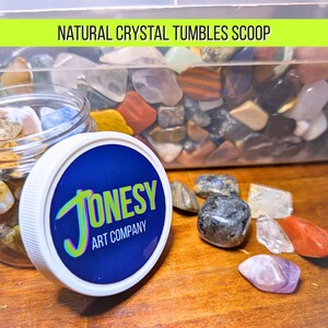 May include: A clear plastic jar filled with assorted polished natural crystal tumbles, featuring a white lid with a blue label that reads JONESY ART COMPANY. A larger clear container of crystals is visible behind the jar, with the text NATURAL CRYSTAL TUMBLES SCOOP displayed above.