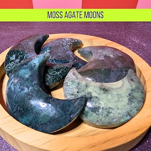 May include: A wooden bowl holds several moss agate moon-shaped stones. The stones display a mix of green, blue, and brown hues. The text "MOSS AGATE MOONS" is displayed in a green rectangle above the bowl.