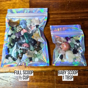 May include: Two iridescent zip-lock bags filled with colorful gemstones. The larger bag, labeled "FULL SCOOP 1/2 CUP", contains a variety of stones. The smaller bag, labeled "BABY SCOOP 1 TBSP", also holds a mix of gems. The background is a dark wood surface.