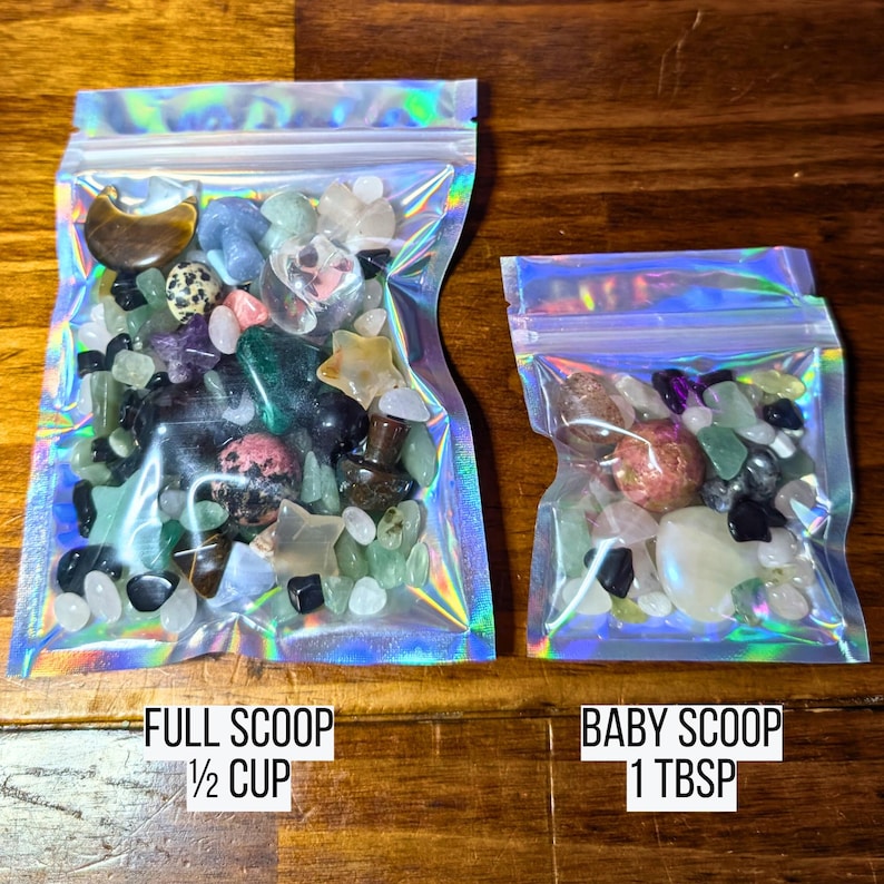 May include: Two iridescent zip-lock bags filled with colorful gemstones. The larger bag, labeled "FULL SCOOP 1/2 CUP", contains a variety of stones. The smaller bag, labeled "BABY SCOOP 1 TBSP", also holds a mix of gems. The background is a dark wood surface.