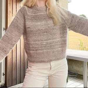 May include: A beige and brown speckled knit sweater with a crew neck and long sleeves. The textured jumper is worn with light-colored trousers. The sweater's design features a horizontal striped pattern.