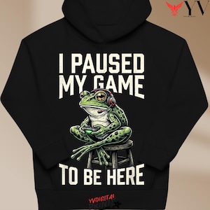 Puede incluir: Sudadera con capucha negra con el texto "I PAUSED MY GAME TO BE HERE". El gráfico presenta una rana verde con auriculares y un mando de videojuegos, sentada en un taburete. La rana tiene manchas negras y ojos amarillos.