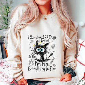 May include: Beige sweatshirt with the text "I Survived 67 Days of School". The graphic features a distressed black cat with large blue eyes, surrounded by school-themed doodles. The text "It's Fine, I'm Fine, Everything Is Fine" is also included.