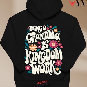 May include: Black hoodie with the text "Being a Grandma is Kingdom Work" in a retro font, surrounded by colorful flowers. The hoodie has a drawstring hood and a kangaroo pocket.