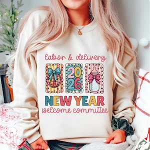 Op de afbeelding: Crèmekleurige sweatshirt met de tekst "labor & delivery" boven drie kleurrijke illustraties. De illustraties omvatten een wereldbol met een strik, de cijfers "2026" en een babyfles met een strik. De tekst "NEW YEAR welcome committee" staat eronder.