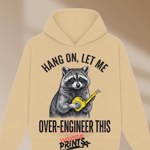 May include: A beige hooded sweatshirt featuring a graphic of a raccoon holding a yellow measuring tape. The text above the raccoon reads HANG ON, LET ME and the text below reads OVER-ENGINEER THIS. The logo YVDIGITAL PRINTS is at the bottom.