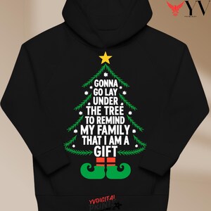 May include: Black hooded sweatshirt with a Christmas tree design. The green tree includes the text "GONNA GO LAY UNDER THE TREE TO REMIND MY FAMILY THAT I AM A GIFT". The tree is topped with a yellow star and elf feet at the bottom.