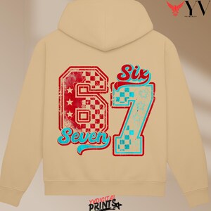 May include: A beige hooded sweatshirt with a large graphic on the back. The graphic features the number "67" in red and blue, with the words "Six" and "Seven" in a stylized font. The design has a distressed, vintage appearance.