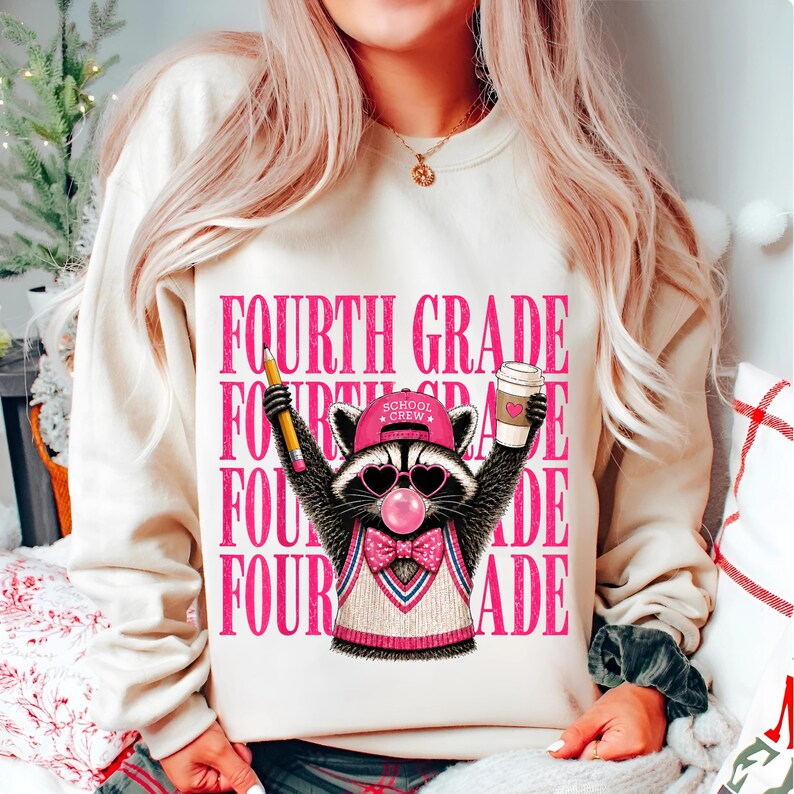 May include: A cream-colored crewneck sweatshirt featuring a graphic of a raccoon wearing a pink cap, heart-shaped sunglasses, and a bow tie. The raccoon holds a pencil and a coffee cup. The text "FOURTH GRADE" is repeated in pink behind it.