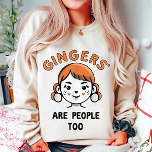 Può includere: Felpa color crema con la scritta "Gingers are People Too" in arancione e nero. La grafica presenta un'illustrazione a cartoni animati di una persona con capelli rossi e lentiggini. La felpa ha maniche lunghe e girocollo.