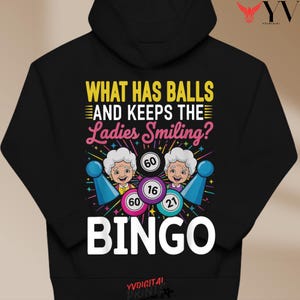 May include: Black hoodie with a colorful bingo-themed graphic. The design includes the text "WHAT HAS BALLS AND KEEPS THE Ladies Smiling?" and "BINGO", along with bingo balls and smiling cartoon characters. The design is printed on the front.