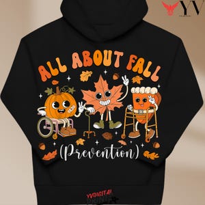 Puede incluir: Una sudadera negra con capucha que presenta un gráfico con el texto ALL ABOUT FALL (Prevention). El diseño incluye una calabaza en silla de ruedas, una hoja de arce con bastón y un trozo de tarta de calabaza con andador, entre hojas.