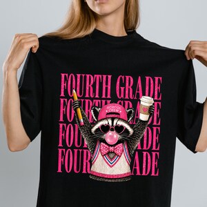 May include: A black t-shirt featuring a graphic of a raccoon wearing a pink cap with the text SCHOOL CREW, heart-shaped sunglasses, a pink bubble, and a bow tie. The raccoon holds a yellow pencil and a coffee cup. The text FOURTH GRADE repeats.