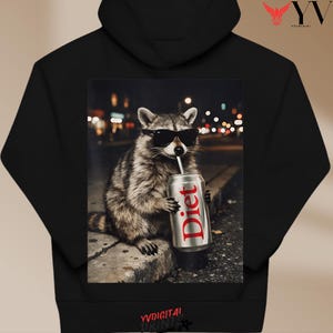 May include: Black hoodie featuring a raccoon wearing sunglasses and drinking from a Diet soda can. The raccoon is sitting on a curb with a blurred city background. The word "Diet" is printed on the can in red.