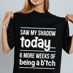 May include: Black t-shirt with white text that reads "SAW MY SHADOW today... 6 MORE WEEKS of being a b*tch". The shirt is a crew neck style.