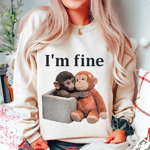 May include: Cream-colored sweatshirt with the text "I'm fine" in black. The graphic features two monkey plush toys, one brown and one gray, hugging inside a gray rectangular container. The sweatshirt is a casual style.