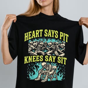 Puede incluir: Camiseta negra con un diseño gráfico que presenta esqueletos y el texto "HEART SAYS PIT KNEES SAY SIT" en amarillo. El diseño incluye una multitud de esqueletos bailando y un esqueleto sentado.