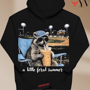 Può includere: Felpa con cappuccio nera con un procione con occhiali da sole e un berretto da baseball, che tiene in mano un caffè freddo. Il procione è seduto su una sedia pieghevole blu con un campo da baseball sullo sfondo. Il testo sulla felpa con cappuccio recita "a little feral summer".