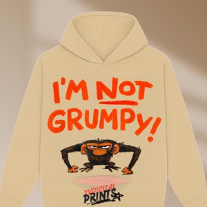 Puede incluir: Sudadera beige con el texto naranja "I'M NOT GRUMPY!". Debajo, una ilustración de un mono gruñón. La sudadera también muestra el texto "YVDIGITAL PRINTS" en la parte inferior.