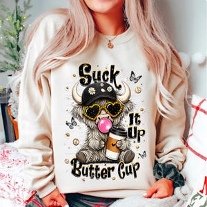 May include: A cream-colored crewneck sweatshirt featuring a graphic of a fluffy highland cow wearing a black baseball cap and heart-shaped sunglasses, blowing a pink bubble, and holding a coffee cup with the text Suck It Up Butter Cup.