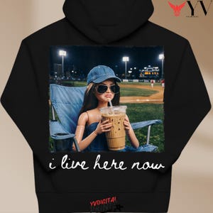 Op de afbeelding: Zwarte hoodie met een afbeelding van een pop met een zonnebril en een spijkerbroekpet, die een ijskoffie vasthoudt. De pop zit in een blauwe klapstoel met een honkbalveld op de achtergrond. De tekst "i live here now" staat eronder.